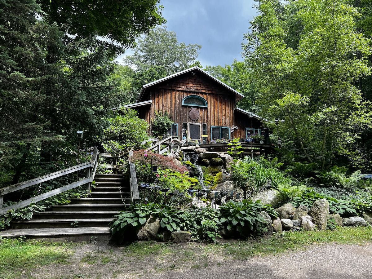 A cabin like home with a water fountain in front of it, all covered in green plants, turned into a pottery store