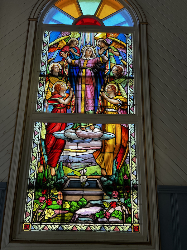 Large stained glass piece featuring a woman (presumably Mother Mary) being ascended from a coffin into heaven by angels