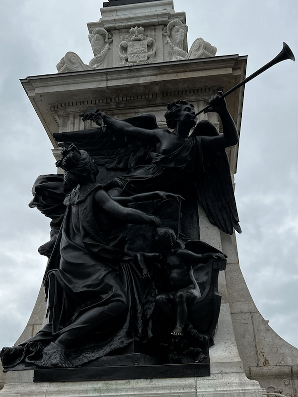 A statue in Trois-Rivières of a mother with some sort of crown, caring for a child, looking up at a passing by angel blowing a horn